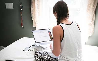 Student working on a bed with laptop, facing away from the camera