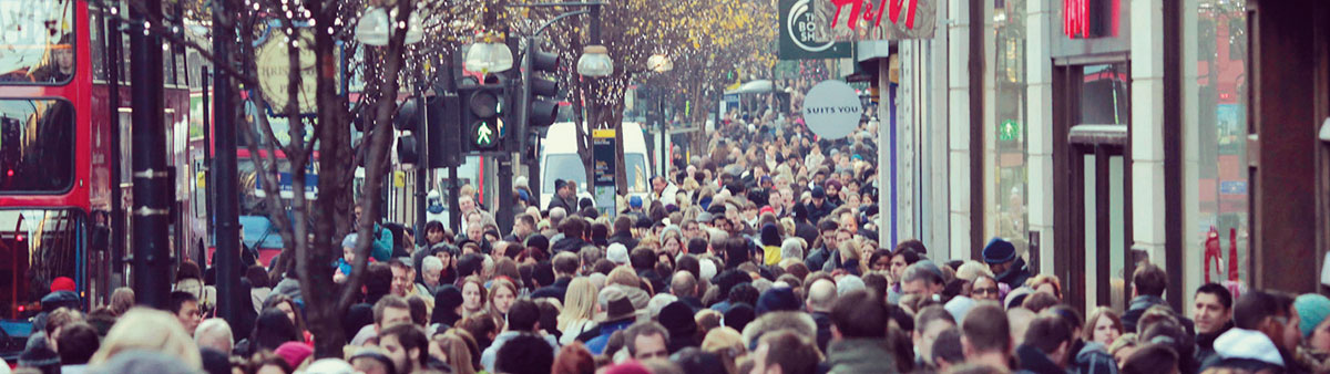 A busy street in London packed with pedestrians. A red double decker bus at one side and shop fronts to the other.